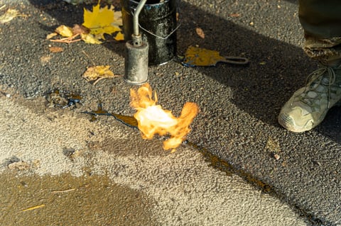 Worker repairing asphalt cracks using a fiery flame gas burner to heat the surface before applying mastic sealant