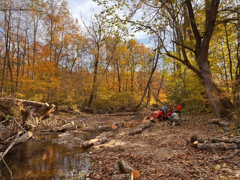 Autumn forest clearing with fallen logs, small stream, and red ATV near large trees with golden foliage