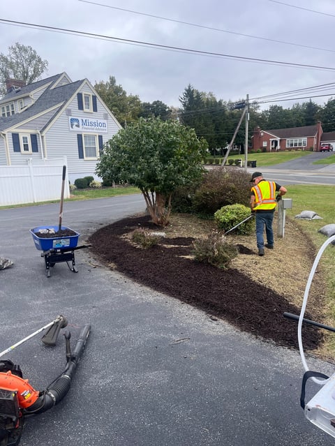 Worker in yellow safety vest spreads mulch around landscape bed near white mission building