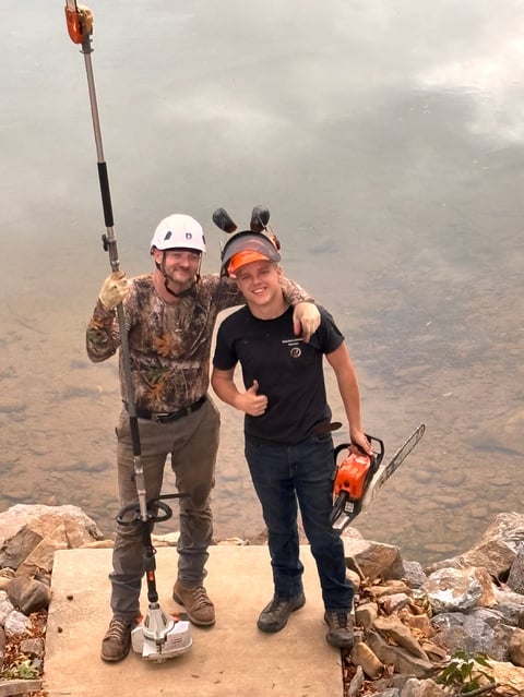 Two workers on a high cliff edge holding forestry tools, wearing safety helmets and protective gear overlooking a mountainous landscape