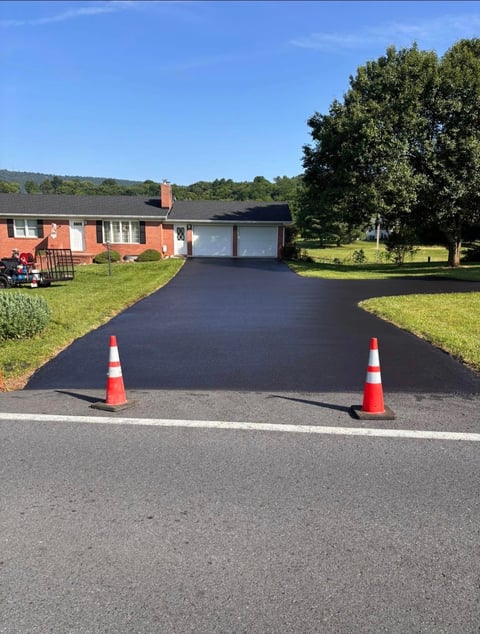 Newly paved driveway with red and white traffic cones in front of a brick ranch house with green lawn
