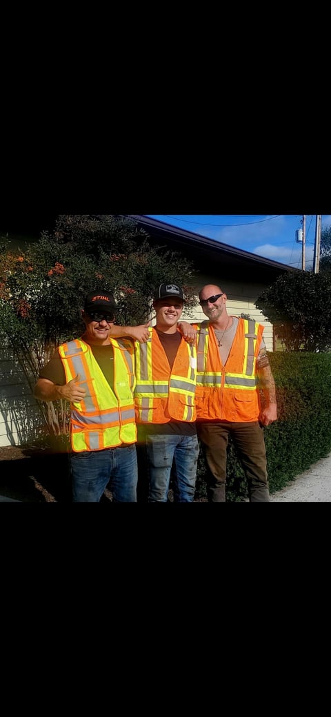 Three men wearing safety vests standing together in front of a house with hedges and blue sky