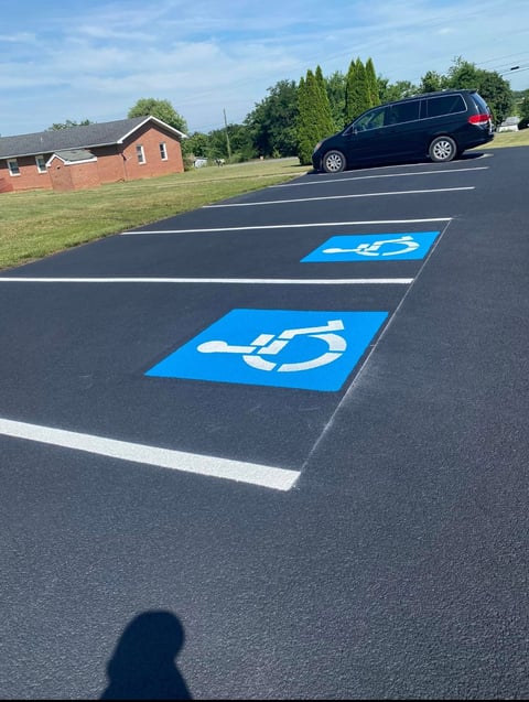 Two blue accessible parking spaces marked with wheelchair symbols on an asphalt lot near a red brick building and parked minivan.