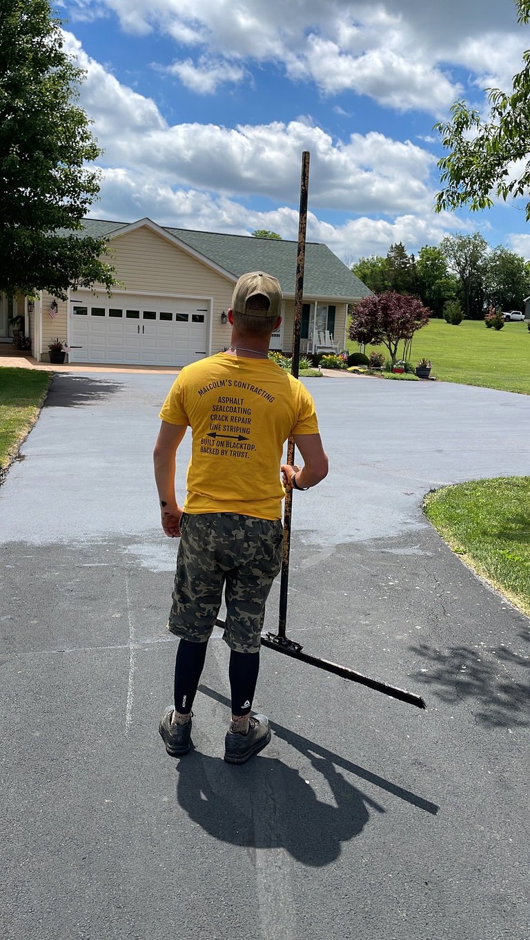 Person in yellow shirt carrying tall wooden pole on residential driveway with house and garage in background