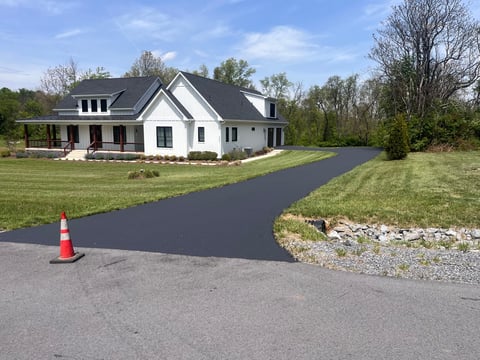 New asphalt driveway being paved in front of a white farmhouse with black roof and manicured lawn