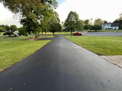 Long asphalt driveway leading to a white two-story house, flanked by large trees and manicured green lawns