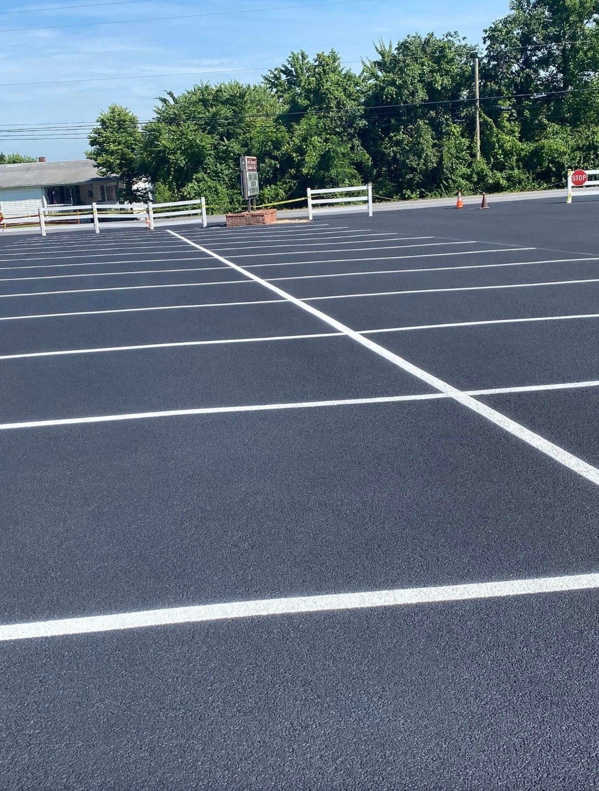 Empty parking lot with white line markings, white fence, and green trees in background under clear blue sky