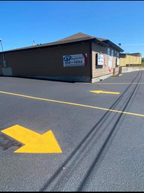 Black restaurant building with yellow arrow painted on asphalt parking lot pointing toward entrance under clear blue sky