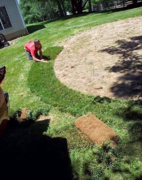 Person in pink shirt seeding or working on lawn areas in a residential yard with green grass and bare soil patches