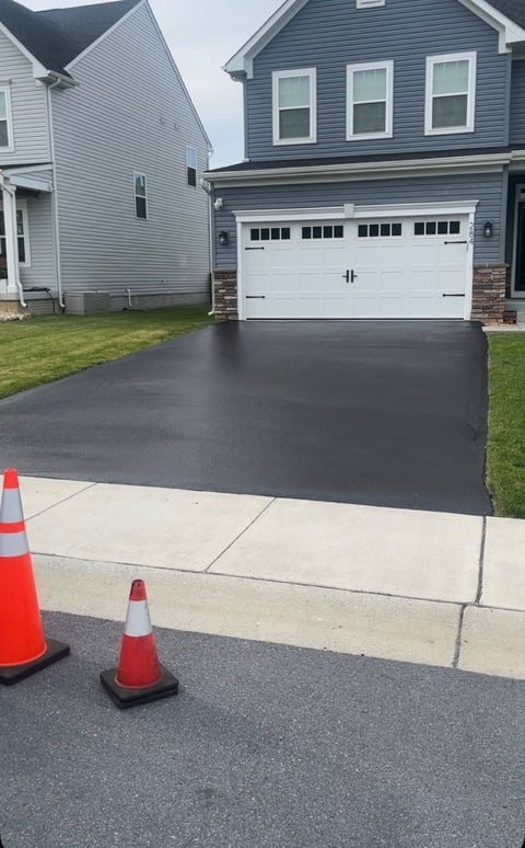 Newly paved black asphalt driveway in front of a two-story gray house with white garage door, marked with traffic cones