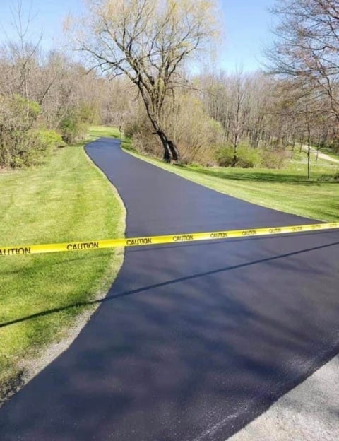 Newly paved curved asphalt driveway with yellow caution tape, bordered by green lawn and bare spring trees