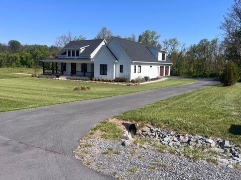 Modern white farmhouse with dark roof on manicured lawn and paved driveway surrounded by trees