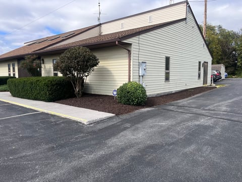 Modern single-story building with cream-colored siding, angled roof, brick foundation, manicured landscaping, and asphalt parking area